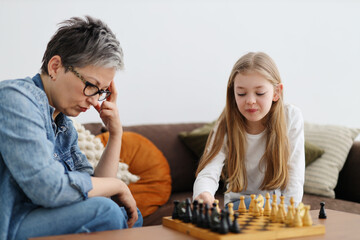 Grandmother plays chess with a girl in a home interior.