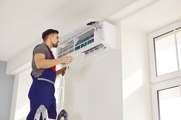 Male technician in uniform repairing air conditioner with screwdriver. Air conditioning master standing on stepladder installing, cleaning or repairing conditioner in apartment