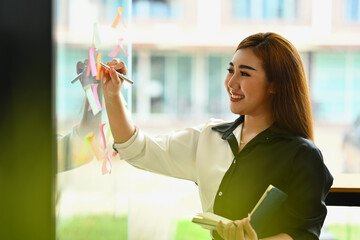 Smiling businesswoman standing near glass wall with colorful sticky papers, working on corporate project strategy in office