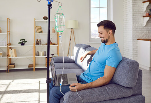 Young man sitting on sofa and having online video call or webinar using laptop at home while receiving IV drip infusion and vitamin therapy in his blood. Male person receiving injection therapy.
