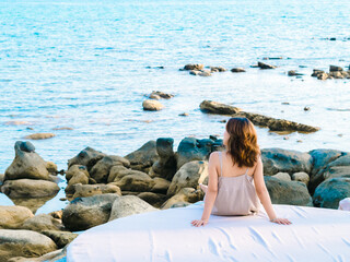 Back of Asian woman in casual dress sitting armrest on cushion near the rocks at coastline, beach in summer vibes. Lonely female relaxing at the sea and looking far away. Holiday travel vacation.
