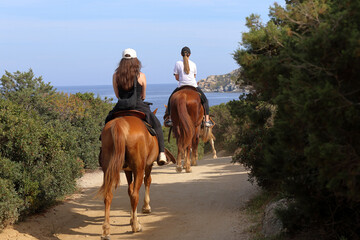 Young girls on horses ride an empty road towards the sea on a beautiful sunny day - 614700429