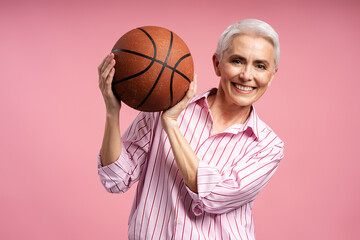 Portrait of beautiful smiling senior gray haired woman holding ball playing basketball game isolated on pink background. Healthy lifestyle concept