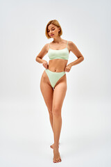 Full length of barefoot and blonde young woman in light green lingerie touching panties while standing and posing on grey background, self-acceptance and body positive concept