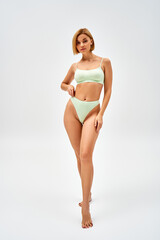 Full length of confident young blonde woman in light green lingerie looking at camera while standing and posing on grey background, self-acceptance and body positive concept