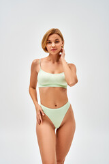 Fototapeta premium Pretty young blonde woman with natural makeup in light green lingerie touching hip while standing and looking at camera isolated on grey, self-acceptance and body positive concept