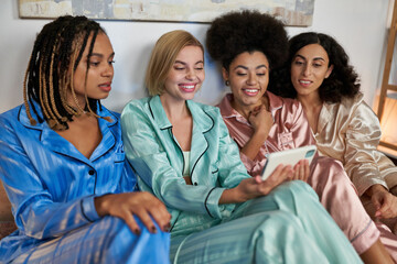 Smiling blonde woman using and holding smartphone near multiethnic girlfriends in colorful pajama during pajama party at home, bonding time in comfortable sleepwear, slumber party
