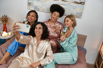 High angle view of multiracial woman in pajama taking selfie with cheerful multiethnic girlfriends with wine and sitting on bed at home, bonding time in comfortable sleepwear, slumber party