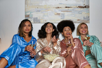 Smiling multiracial women in pajama watching tv and holding popcorn and glasses of wine while...