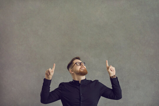 Smiling man looks up and points his index fingers at the free space above his head, offering something to choose from. People and promotion concept. Place for text. Gray background.