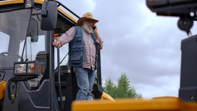 Wide shot bearded senior farmer in straw hat talking on phone in slow motion standing at tractor. Portrait of confident Caucasian expert man at harvester outdoors