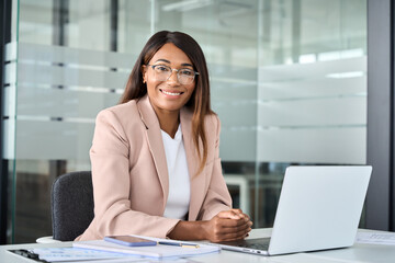 Young happy professional African American business woman wearing suit eyeglasses working on laptop in office sitting at desk looking at camera, female company manager executive portrait at workplace.