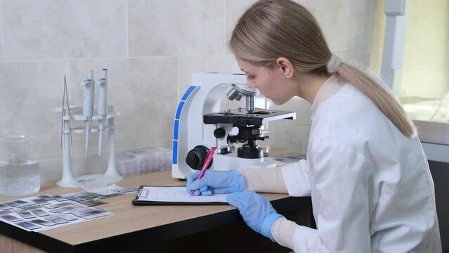 a female laboratory assistant studies microorganisms by looking through a microscope and writes the readings in a log.