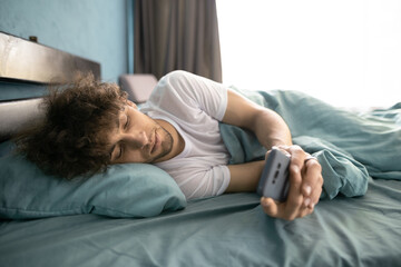 Arabic male awake lying in comfortable bed checking mail on smartphone, happy guy wake up in morning using cellphone reads message or text - Powered by Adobe