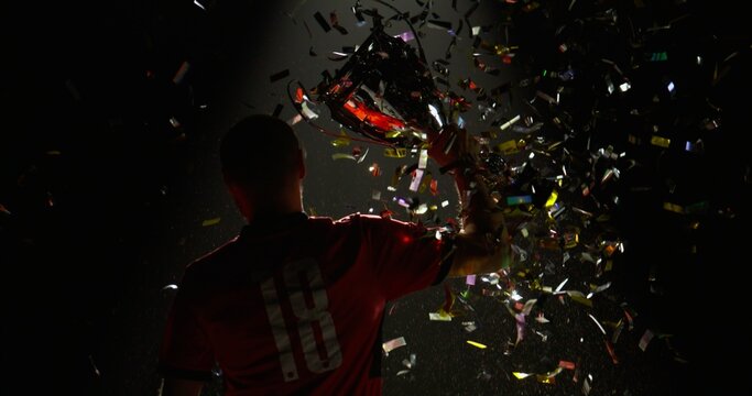 Silhouette of Caucasian male soccer football player raising a trophy above head against bright light and falling confetti. Super slow motion, shot on RED cinema camera