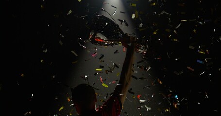 Silhouette of Caucasian male soccer football player raising a trophy above head against bright light and falling confetti. Super slow motion, shot on RED cinema camera