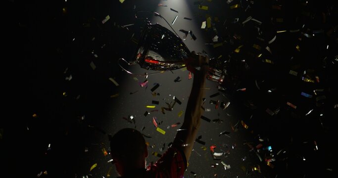 Silhouette of Caucasian male soccer football player raising a trophy above head against bright light and falling confetti. Super slow motion, shot on RED cinema camera