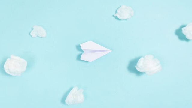 White paper airplane flies through white clouds. Blue background. Looped movement. Travel and transportation concept. Stop motion animation. Copy space.