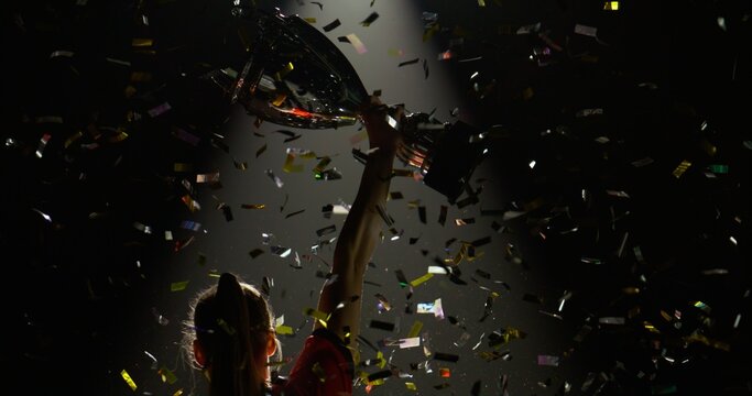 Silhouette of Caucasian woman female soccer football player raising a trophy above head against bright light and falling confetti. Super slow motion, shot on RED cinema camera