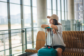 Young cheerful dreamy woman sitting at the airport in the waiting area for departures with a suitcase. Summer travel concept