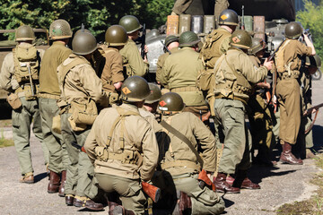 Fototapeta premium Historical reconstruction. An American infantry soldiers from the World War II prepare to attack hidden behind a tank. View from the back.