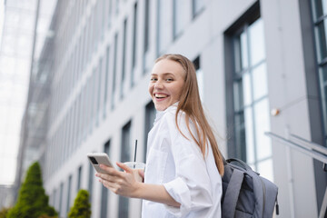 Fototapeta premium Business women style. Woman going to work chatting on phone while going upstairs. Portrait of beautiful smiling female hold take away ice late, reading news on her mobile, look happy, scrolling page.