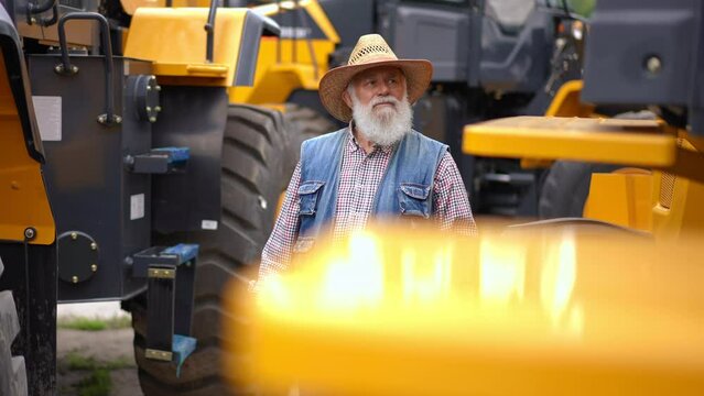 Portrait of confident focused senior man walking between rows of new agricultural vehicles choosing new tractor. Professional expert Caucasian farmer buying cultivator outdoors