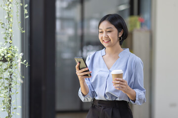 Fototapeta na wymiar Young beautiful Asian woman holding coffee paper cup and looking at smartphone while sitting at workplace. Happy university student girl using mobile phone. Business woman drinking coffee and smiling.