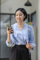 Young beautiful Asian woman holding coffee paper cup and looking at smartphone while sitting at workplace. Happy university student girl using mobile phone. Business woman drinking coffee and smiling.