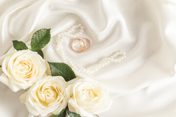 Obraz na płótnie Canvas Two gold wedding rings on a delicate satin fabric background with pearl beads and white roses. A copy of the space. Design layout.