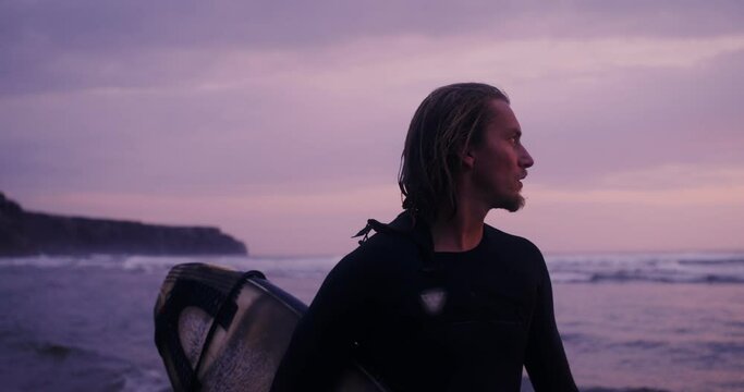 A man in a diving suit with a surfboard in his hands stands on the seashore at sunset