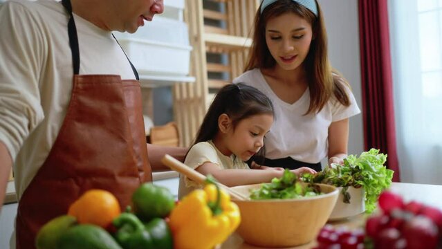 Young asian family cooking food in kitchen.Cute little girl and her beautiful parents are making salad and smiling while cooking in kitchen at home.Happy family in the kitchen concept.