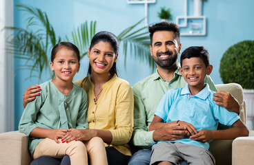 Happy smiling indian couple with sibling kids looking or posing to camera while sitting on sofa at...