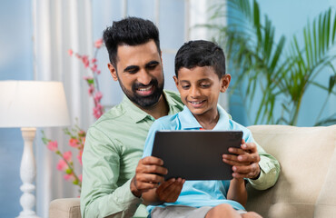 Happy Indian father teaching at home teaching from digital tablet while sitting on sofa - conept of...