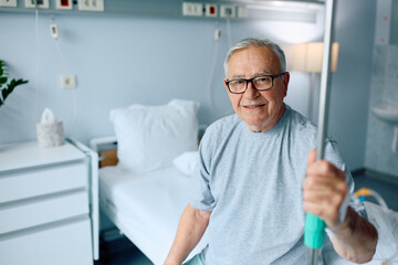 Happy senior man with IV drip sits on hospital bed while looking at camera.