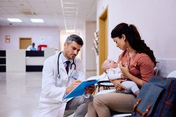 Pediatrician take notes while talking to mother with baby at medical clinic.