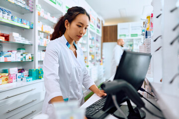 Young Chinese pharmacist working on desktop PC in drugstore.