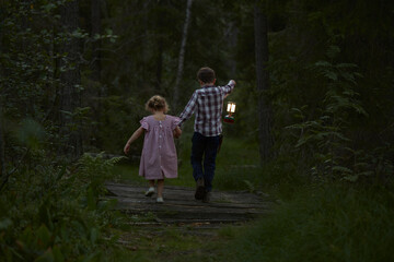 Brother and sister walking with lantern over footbridge in woods - obrazy, fototapety, plakaty