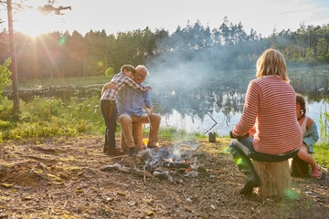 Grandparents and grandchildren hugging at campfire at sunny lakeside