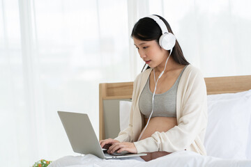 Pregnant Asian mom sitting on her bed working on her laptop and wearing headphones. Drinking orange juice makes the body healthy. Work from home with Wi-Fi technology. concept of motherhood