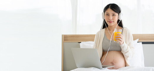 Pregnant Asian mom sitting on her bed working on her laptop and wearing headphones. Drinking orange juice makes the body healthy. Work from home with Wi-Fi technology. concept of motherhood