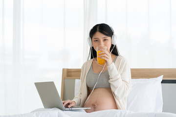 Pregnant Asian mom sitting on her bed working on her laptop and wearing headphones. Drinking orange juice makes the body healthy. Work from home with Wi-Fi technology. concept of motherhood
