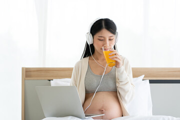 Pregnant Asian mom sitting on her bed working on her laptop and wearing headphones. Drinking orange juice makes the body healthy. Work from home with Wi-Fi technology. concept of motherhood