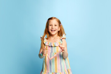 Portrait of beautiful, happy, little girl, child in multicolored striped dress posing with smile and laugh against blue studio background