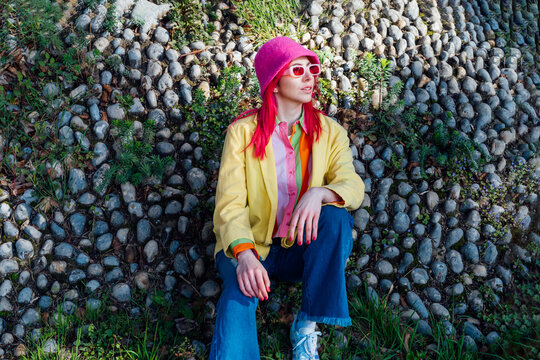 Urban spring street fashion look. Vanilla Girl. Kawaii vibes. Candy colors design. Bucket hat trends. Young woman with pink hair and sunglasses in multicolor outfit on the stones and plants background