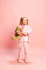Full-length portrait of cute little girl, child standing with many books and bear toy against pink studio background. Education and game