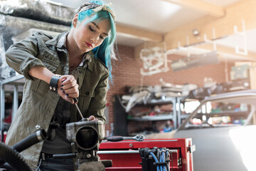 Young female mechanic blue hair repairing part in auto repair shop - obrazy, fototapety, plakaty