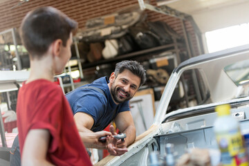 Smiling father taking tool from son in auto repair shop - obrazy, fototapety, plakaty