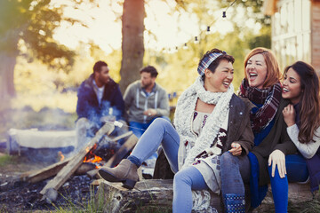 Female friends laughing and roasting marshmallows at campfire - obrazy, fototapety, plakaty