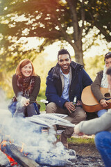 Smiling friends roasting marshmallows and drinking beer at campfire